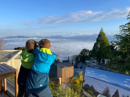 Zwei Kinder stehen am Rand einer Terrasse und blicken auf die schneebedeckten Berge über einer Nebeldecke.