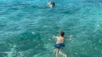 Zwei Kinder schwimmen fröhlich im klaren, türkisfarbenen Wasser unter einem strahlend blauen Himmel.