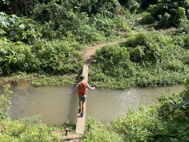 Ein Wanderer balanciert auf einem schmalen Steg über einen ruhigen Fluss, umgeben von üppigem, grünem Dschungel.