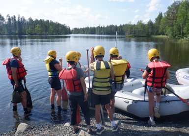 Eine Gruppe von sechs Personen in Schwimmwesten und Helmen steht am Ufer eines ruhigen Sees, bereit zum Rafting.