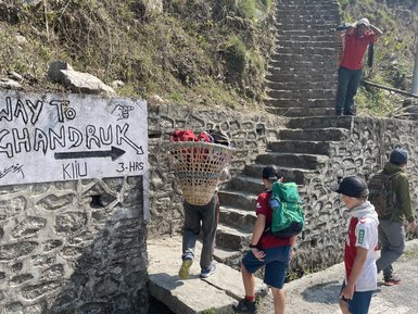 Eine Gruppe von Kindern steht an einer steinernen Treppe, während ein Erwachsener mit einem Korb auf dem Rücken vorbeigeht.