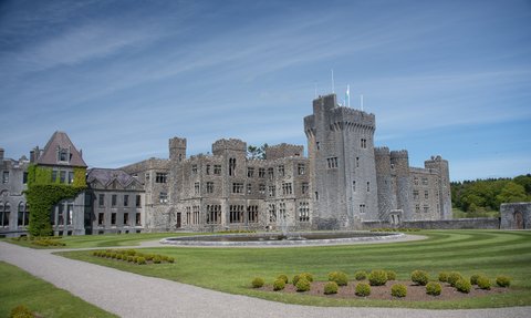 Ein beeindruckendes Schloss mit grauer Steinfassade und Türmen, umgeben von gepflegten Rasenflächen und Sträuchern.