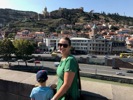 Eine Frau in einem grünen Kleid lächelt, während sie mit einem Jungen an einer Mauer steht und die Stadt Tiflis betrachtet.