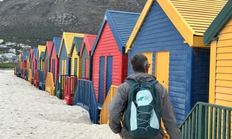 Ein Mann mit einem Rucksack geht an bunten Strandhütten entlang, die in verschiedenen Farben gestrichen sind.