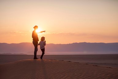Eine Mutter und ihr Kind stehen auf einer Sanddüne, als die Sonne hinter ihnen aufgeht und die Landschaft in warmen Tönen erstrahlt.