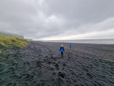 Zwei Kinder in blauen Jacken laufen über einen schwarzen Sandstrand, umgeben von grauen Wolken und sanften Hügeln.