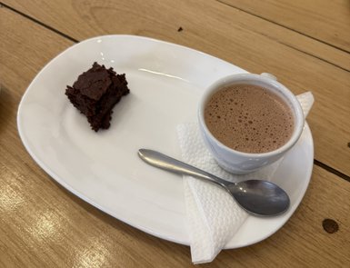 Ein Stück schokoladiger Kuchen liegt auf einem weißen Teller neben einer Tasse heißer Schokolade und einem Löffel.