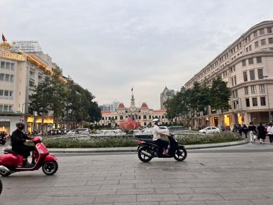 Eine belebte Straße in Saigon mit Motorrädern und einem großen Brunnen im Vordergrund, umgeben von historischen Gebäuden.