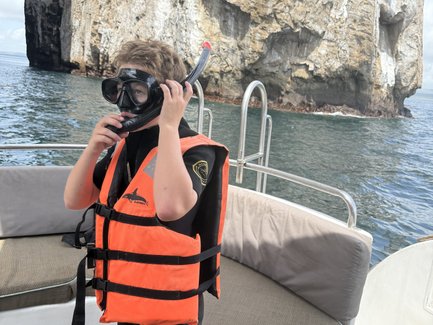 Ein Junge mit einer Schnorchelmaske und orangefarbener Schwimmweste steht auf einem Boot vor einer beeindruckenden Felswand.