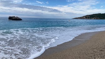 Ein ruhiger Strand mit sanften Wellen, die an den feinen Sand spülen, während ein Felsen im klaren Wasser sichtbar ist.