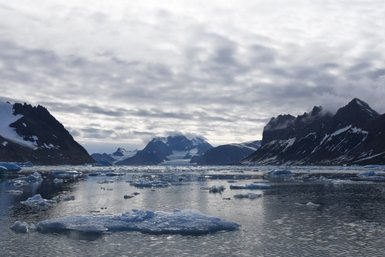 Eine ruhige, eisbedeckte Wasserfläche spiegelt die majestätischen Berge und den bewölkten Himmel in Hornsund wider.