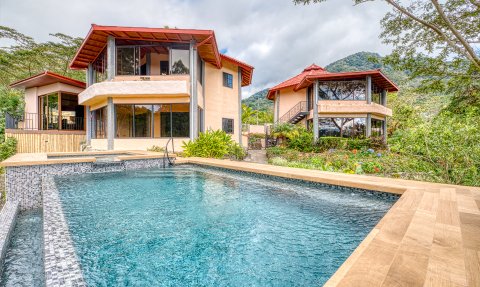 Zwei moderne Häuser mit roten Dächern stehen neben einem glitzernden Pool, umgeben von üppigem Grün und Bergen im Hintergrund.