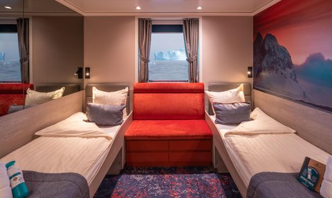 Ein gemütliches Kabineninnere mit zwei Betten, einem roten Sofa und einem Fenster, das auf Eisberge im Ozean blickt.