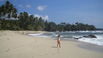 Ein Kind läuft barfuß am Strand entlang, während die Wellen sanft ans Ufer schlagen und Palmen im Hintergrund stehen.