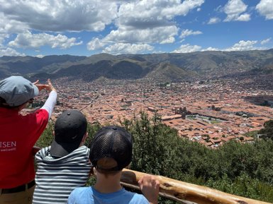 Eine Person zeigt auf die Stadt Cusco, während zwei Kinder fasziniert auf die Aussicht blicken, umgeben von Bergen.
