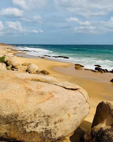 Eine ruhige Küstenlandschaft mit großen Felsen, goldenem Sand und sanften Wellen, die an den Strand rollen.