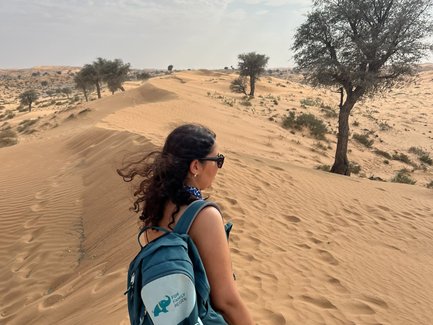 Eine Frau mit Sonnenbrille und Rucksack wandert durch die goldenen Sanddünen unter einem bewölkten Himmel.