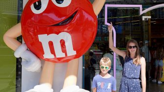 Ein großer, roter M&M's Charakter steht vor einem Geschäft, während ein Junge und ein Mädchen lächeln und posieren.