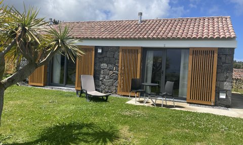 Ein modernes, steinernes Ferienhaus mit Holzjalousien, umgeben von grünem Rasen und einer Palme im Vordergrund.