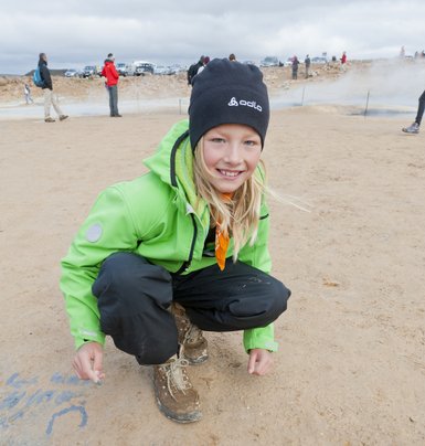 Ein lächelndes Kind in einer grünen Jacke kniet auf dem Boden und schreibt mit einem Stift in den Sand.