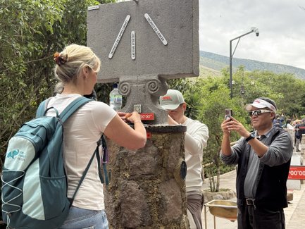 Eine Frau mit einem Rucksack interagiert mit einem Steinsäule, während ein Mann sie mit einem Smartphone filmt.