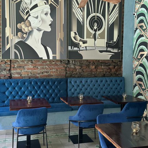 Ein stilvolles Café mit blauen Samtsofas und eleganten Holztischen, umgeben von kunstvollen Wandmalereien.