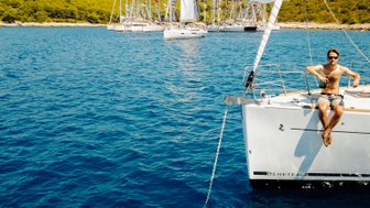 Ein junger Mann sitzt entspannt auf der Reling einer Segelyacht und genießt die Sonne auf dem blauen Wasser.