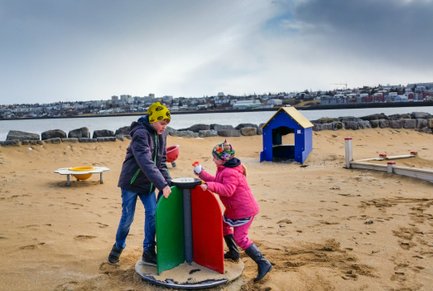 Zwei Kinder spielen auf einem Sandspielplatz, während im Hintergrund eine Stadtlandschaft und ein ruhiger Wasserbereich sichtbar sind.