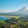 Ein majestätischer Vulkan erhebt sich über eine grüne Landschaft, umgeben von einem klaren blauen See und sanften Hügeln.