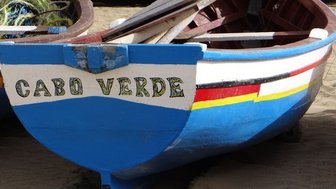 Ein buntes Fischerboot mit der Aufschrift 'Cabo Verde' steht am Strand, umgeben von weiteren Booten und Seilmaterial.