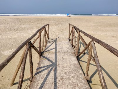 Eine Holzbrücke führt über den Sandstrand zu den sanften Wellen des Meeres, unter einem klaren blauen Himmel.