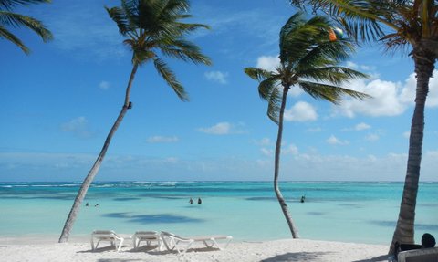 Ein tropischer Strand mit weißen Liegen, umgeben von Palmen und klarem, türkisfarbenem Wasser unter einem strahlend blauen Himmel.