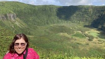 Eine lächelnde Frau in einem pinken Hemd steht vor einer beeindruckenden, grünen Vulkanlandschaft unter blauem Himmel.