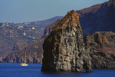 Ein majestätischer Felsen erhebt sich aus dem blauen Wasser, während ein Segelboot in der Nähe sanft schaukelt.