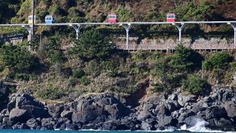 Eine farbenfrohe Seilbahn fährt über eine grüne Küstenlandschaft, während Wellen sanft gegen die Felsen schlagen.