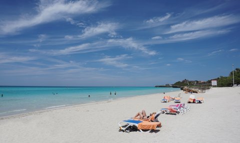 Ein sonniger Strand mit weißen Sandflächen, Liegen und Menschen, die im klaren Wasser schwimmen und sich entspannen.