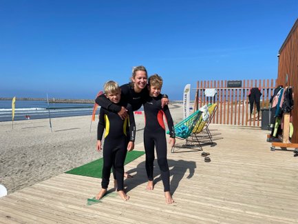 Eine Frau umarmt zwei Kinder in Neoprenanzügen am Strand, bereit zum Surfen, mit Wellen im Hintergrund.
