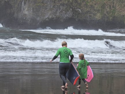 Eine Frau und ein Mädchen in grünen Neoprenanzügen gehen mit Bodyboards am Strand entlang, während die Wellen brechen.