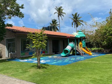Ein farbenfroher Spielplatz mit Rutschen und einem Spielhaus, umgeben von grünem Rasen und Palmen unter einem blauen Himmel.
