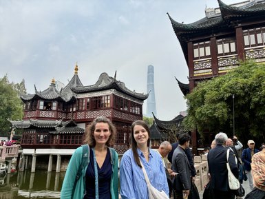Zwei Frauen lächeln vor traditionellen Holzgebäuden in Shanghai, während moderne Wolkenkratzer im Hintergrund sichtbar sind.