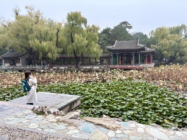 Eine Frau in einem langen, hellen Mantel steht an einem Steg und betrachtet die ruhige Wasseroberfläche eines Sees.
