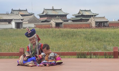 Zwei Kinder in traditioneller mongolischer Kleidung sitzen auf einem Holzdeck vor dem Erdene Zuu Kloster in der Natur.