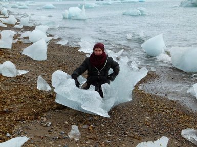 Eine Person sitzt auf einem großen Eisblock am Ufer, umgeben von kleineren Eisstücken und einem ruhigen Wasserhorizont.