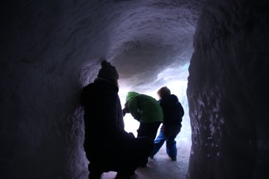 Eine Person sitzt in einer Eishöhle und beobachtet zwei Kinder, die neugierig nach draußen schauen.
