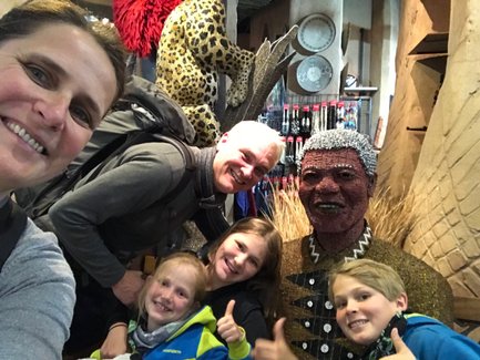 Eine fröhliche Familie posiert vor einer kunstvollen Statue, die einen berühmten Mann darstellt, umgeben von bunten Dekorationen.