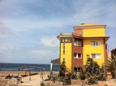 Ein farbenfrohes Gebäude mit der Aufschrift 'Residencial Sol Point' steht am Strand, umgeben von Pflanzen und Meer.