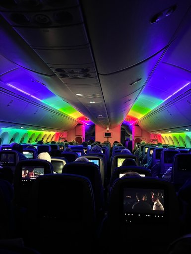 Der Innenraum eines Flugzeugs ist in bunten Lichtern beleuchtet, während Passagiere Filme auf Bildschirmen ansehen.
