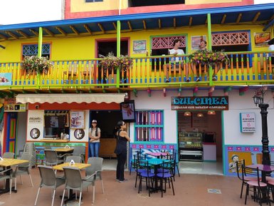 Bunte Fassade eines Cafés mit blühenden Pflanzen und zwei Personen auf dem Balkon, die sich unterhalten.