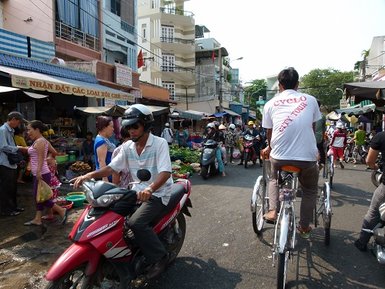 Eine belebte Straße in Saigon, gesäumt von Ständen mit frischen Lebensmitteln und Menschen, die geschäftig unterwegs sind.