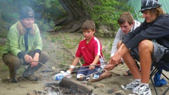 Eine Gruppe von vier Jugendlichen sitzt um ein Lagerfeuer und röstet Marshmallows auf langen Stöcken.